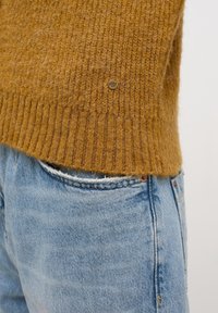 Senapsgul stickad tröja med ribbad nederkant, som har en liten metallisk logotypdetalj. Tillsammans med blekta blå jeans.