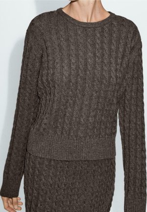Pull à manches longues en maille torsadée marron foncé assorti à une jupe en maille torsadée portée par une personne sur un fond uni.