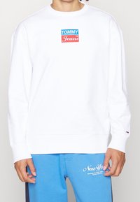 Bílá mikina s modro-červeným logem "TOMMY Jeans". Dlouhé rukávy a design s kulatým výstřihem. Nosit se s modrými tepláky.