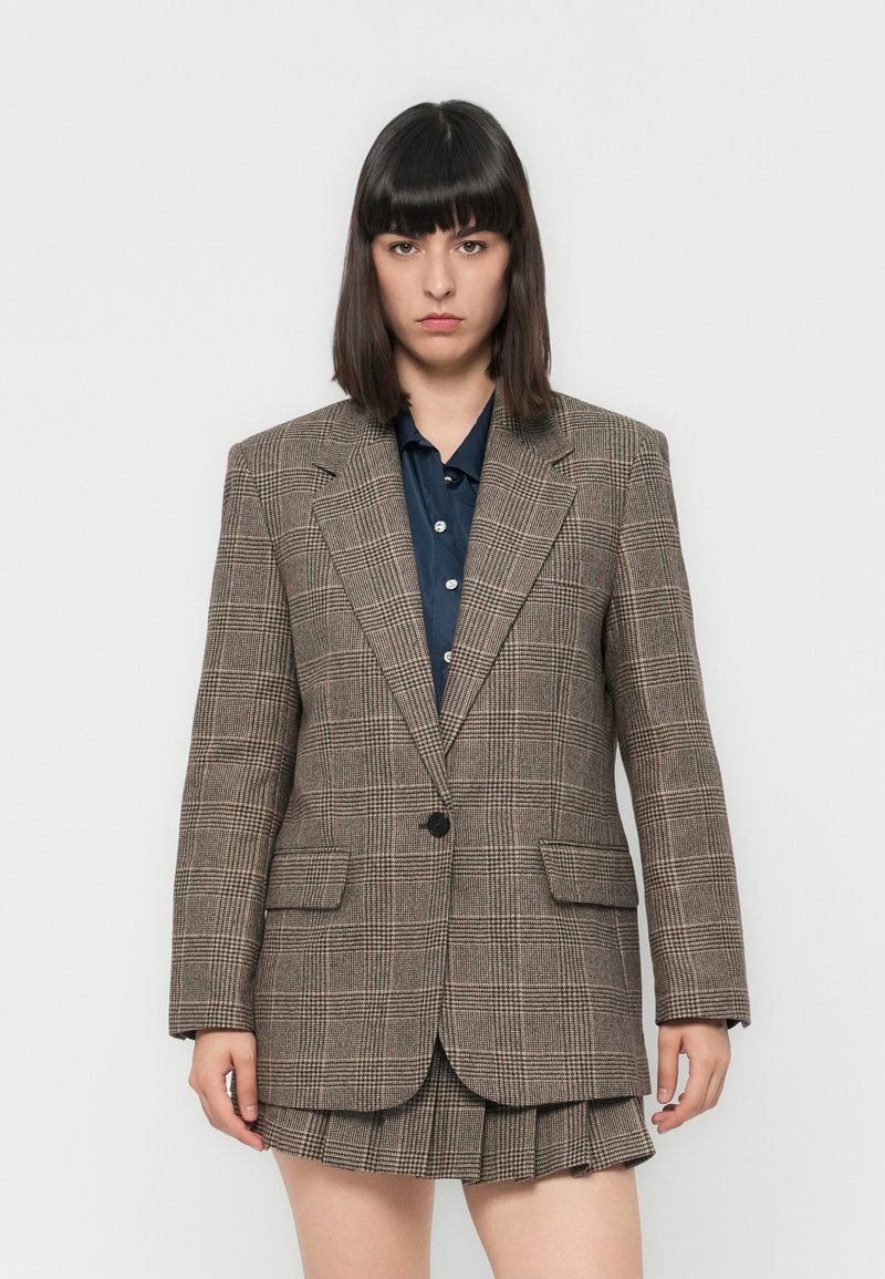 Blazer a quadri marroni con un solo bottone, due tasche frontali, rever affilati, abbinato a una camicia blu navy e una gonna plissettata. Texture liscia.