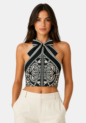 Donna con lunghi capelli castani indossa un crop top allacciato al collo bianco e nero con audaci motivi floreali e pantaloni beige a vita alta, di fronte.