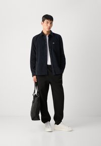 Marine corduroy overhemd met knopen en zak, wit T-shirt eronder, zwarte sweatpants, witte sneakers en een zwarte tote bag.