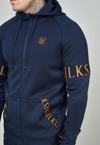 Felpa con zip blu navy con logo dorato sul petto, accenti dorati a motivo a contrasto su maniche e tasche, realizzata in tessuto liscio ed elastico.