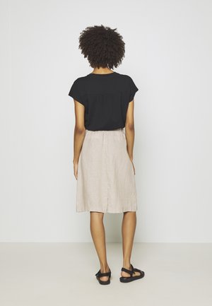 Kvinde med krøllet hår står vendt mod væggen, iført sort kortærmet top, beige knælange nederdel og sorte sandaler på ensfarvet baggrund.