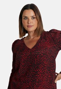 Top s leopardím potiskem v červené a černé barvě, s designem do V, dlouhými rukávy se zúženými detaily na ramenech, vyrobený z měkké a hladké tkaniny.