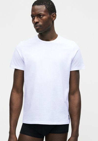 Uomo con capelli corti e ricci che indossa una t-shirt bianca a maniche corte semplice e slip neri, che guarda leggermente verso sinistra.