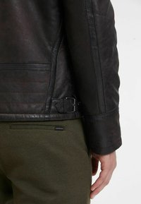 Giacca in pelle nera con texture trapuntata e dettaglio con fibbia in vita, abbinata a pantaloni verde oliva con un piccolo etichetta del marchio.