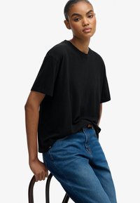 Sort bomulds T-shirt med korte ærmer og rund hals, parret med blå denim jeans. Modellen læner sig op ad en stol.