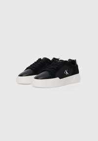 Adidași negri cu un amestec de piele întoarsă și piele lucioasă, având o talpă albă, groasă, și un logo brodat pe lateral.