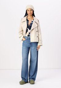 Beige lyhyempi kaksirivinen trench coat sinisen paidan yli, jossa on valkoinen abstrakti printti, yhdistettynä leveälahkeisiin sinisiin farkkuihin ja vihreisiin puukenkäisiin.