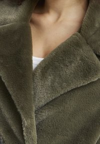 Olivgrön fuskpälsjacka med en mjuk textur, som har en bred krage och en dubbelknäppt design. Nära bild av tygdetaljer.