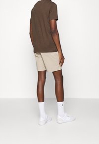 Brun t-shirt med rund hals, beige shorts, vita sneakers och strumpor. Släta material med avslappnad passform. Synlig logotyp på sidan av skon.