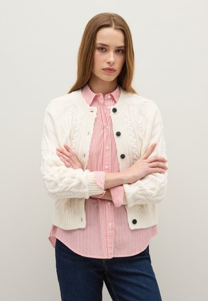 Cremfarbener, kabelstrickartiger Cardigan mit Rundhalsausschnitt, schwarzen Knöpfen, getragen über einem rot-weiß gestreiften Hemd, kombiniert mit dunklen Jeans.