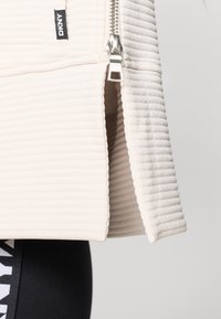 Ανοιχτό καφέ καπιτονέ τζάκετ με φερμουάρ, διαθέτοντας ριγωτή υφή και ετικέτα DKNY. Κοντή σχεδίαση με ίσια γραμμή και ελάχιστες λεπτομέρειες.