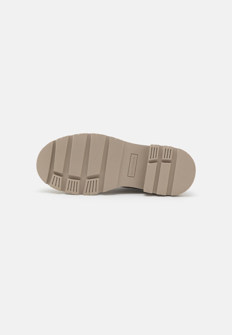 Semelle de chaussure en caoutchouc beige clair, présentant des rainures et des crêtes profondes pour une meilleure adhérence. Surface texturée avec le logo de la marque au centre.