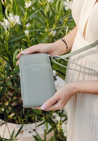 Light green crossbody bag made of synthetic material with a zip closure, embossed logo, and adjustable strap, held against foliage.