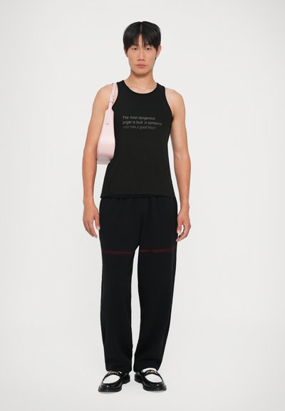 Zwarte tanktop met een geprinte quote, gecombineerd met losse zwarte broek met rode stiksels. Een kleine roze handtas hangt schuin over de schouder.