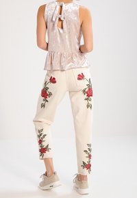 Top in velluto rosa chiaro con peplum e schiena a chiave, pantaloni crema con ricami di rose rosse, abbinati a sneakers grigie.