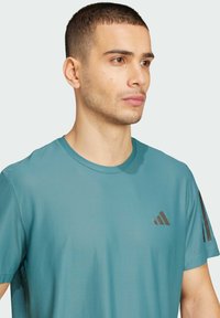 T-shirt de sport à manches courtes teal avec un tissu texturé, encolure ronde et accents noirs rayés sur les épaules. Logo à trois rayures sur la poitrine.