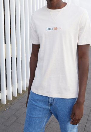 Person wearing a light beige t-shirt with "HILFIGER" text in multicolor embroidery and blue jeans standing by a white slatted fence.