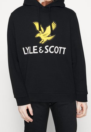 Osoba nosząca czarną bluzę z kapturem z żółtym logo orła i białym napisem "LYLE & SCOTT" na piersi, w połączeniu z czarnymi spodniami.