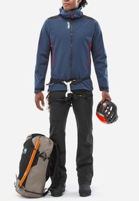 Personne portant une veste bleue et un pantalon de randonnée noirs avec un harnais, tenant un casque d'escalade orange et noir, se tenant à côté d'un sac à dos gris avec une pioche.