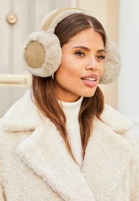 Auriculares de piel sintética en beige y crema, diseño redondeado con textura suave, combinados con un acogedor suéter crema bajo un abrigo texturizado.