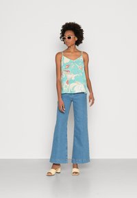 Uma blusa sem mangas de flores em azul-petróleo com alças finas, combinada com jeans flare azul-claro e sandálias de salto bloco bege. Cabelo castanho texturizado.