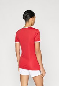 Κόκκινο αθλητικό μπλουζάκι με λευκή επένδυση, κοντά μανίκια και εφαρμοστή σχεδίαση. Συνδυάζεται με λευκά σορτς που έχουν μαύρες ρίγες στα πλαϊνά.