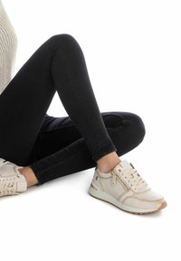 Zapatillas beige con textura suave, diseño con cordones, cremalleras doradas y detalles color marrón, combinadas con jeans negros ajustados.