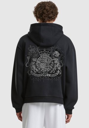 Man wearing black hoodie with detailed crest featuring crowned lion and unicorn holding a shield on the back.