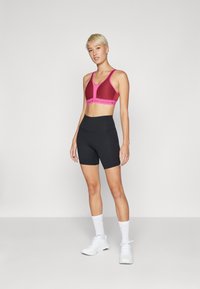 Sujetador deportivo en rosa oscuro con una banda en un rosa más claro como contraste; combinado con pantalones cortos altos negros. Zapatillas y calcetines blancos completan el atuendo.