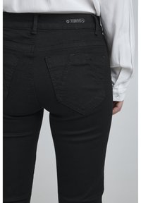 Jeans de mezclilla negra con un diseño ajustado, bolsillos traseros con costura doble y un detalle de botón de metal en la cintura. Textura suave.