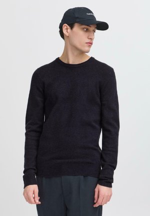 Dunkelblauer Pullover aus weichem Strickstoff, mit Rundhalsausschnitt und langen Ärmeln. Kombiniert mit einer schwarzen Baseballkappe.