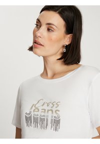 Biała koszulka z krótkimi rękawami, z teksturowanym drukiem graficznym w złotym i czarnym kolorze z napisem "Cross Jeans" o gładkim wykończeniu.