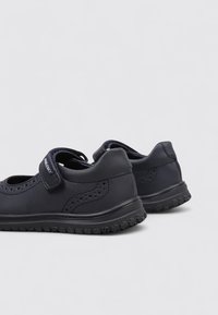 Zapatos tipo Mary Jane para niños negros con correas de Velcro y suelas de goma texturizadas sobre un fondo claro y liso.