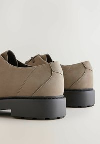 Pantofi din suede bej cu toc mic, vârf rotunjit și talpă din cauciuc negru contrastant. Detalii subtile de cusătură pe partea superioară.