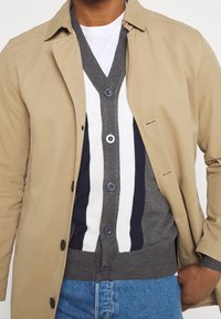 Beige bomullsjacka över en grå, vit och marinrandig cardigan, med knappar och rund halsringning, ihop med ljusblå jeans.