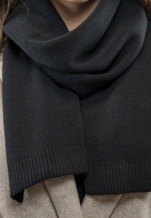 Écharpe noire en tricot enroulée autour du cou, portée sur un manteau beige.