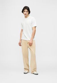 Νέος άνδρας που στέκεται, φορώντας ένα απλό λευκό κοντομάνικο T-shirt, μπεζ παντελόνι με φαρδιά μπατζάκια και λευκά αθλητικά παπούτσια με γκρι σόλες.