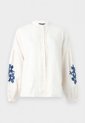 Frau trägt eine weiße Bluse mit blauer Blumenstickerei an den Ärmeln, blaue hoch taillierte weite Hosen und braune Sandalen, steht vor einem schlichten Hintergrund.