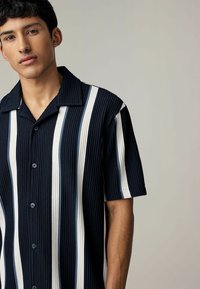 Camisa de manga corta a rayas azul marino y blanca con textura acanalada, cierre de botones en el frente y un suave cuello. Presenta rayas verticales alternas.