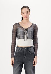 Gekürztes kariertes Oberteil aus transparentem schwarzem Mesh mit langen Ärmeln, Bindeband-Verschluss, über einem weißen, enganliegenden Oberteil getragen. Kombiniert mit hoch taillierter Jeans.