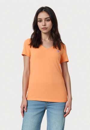 Ung kvinde med langt mørkt hår iført en orange T-shirt med V-udskæring og blå jeans, står foran en ensfarvet lys baggrund.