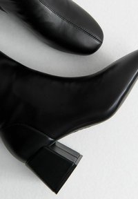 Botas de cuero negras hasta el tobillo con punta afilada y tacón cuadrado. Textura suave con un detalle de costura sutil en la parte frontal.