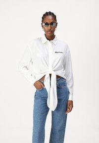 Λευκό πουκάμισο με μακριά μανίκια και κόμπο μπροστά, με λογότυπο στο στήθος. Συνδυασμένο με τζιν μπλε ψηλής μέσης.