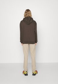 Felpa marrone con cappuccio, abbinata a pantaloni beige chiaro. Il/La indossatore/trice ha i capelli ricci e indossa sneakers nere e gialle.