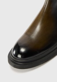 Bottine en cuir avec un capuchon brillant noir, un dessus en cuir marron lisse et une semelle en caoutchouc texturée. Le design courbé améliore l'ajustement.