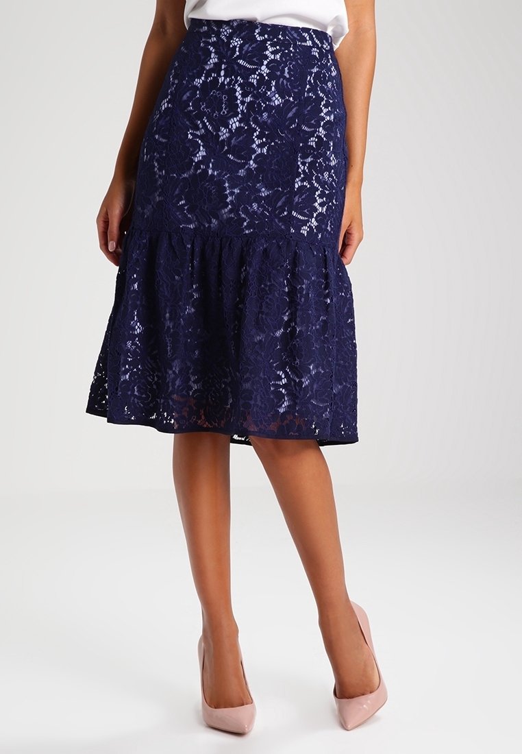 Jupe midi en dentelle bleu marine avec motif floral, ourlet évasé, portée avec un haut blanc et des talons hauts rose pâle sur un fond neutre.