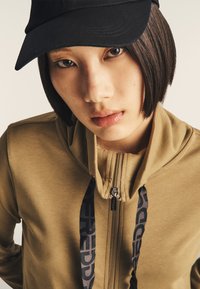 Sudadera con cremallera color caqui y cordones negros con logo; textura suave; combinada con una gorra negra; primer plano que resalta las características faciales.